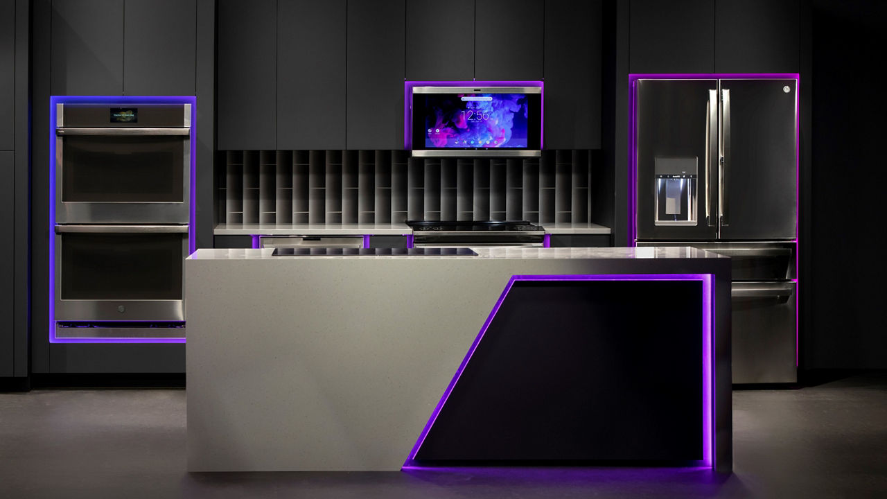 a futuristic kitchen with dark gray cabinets and floors, white quartz countertops, and LED lighting framing all of the GE appliances. 