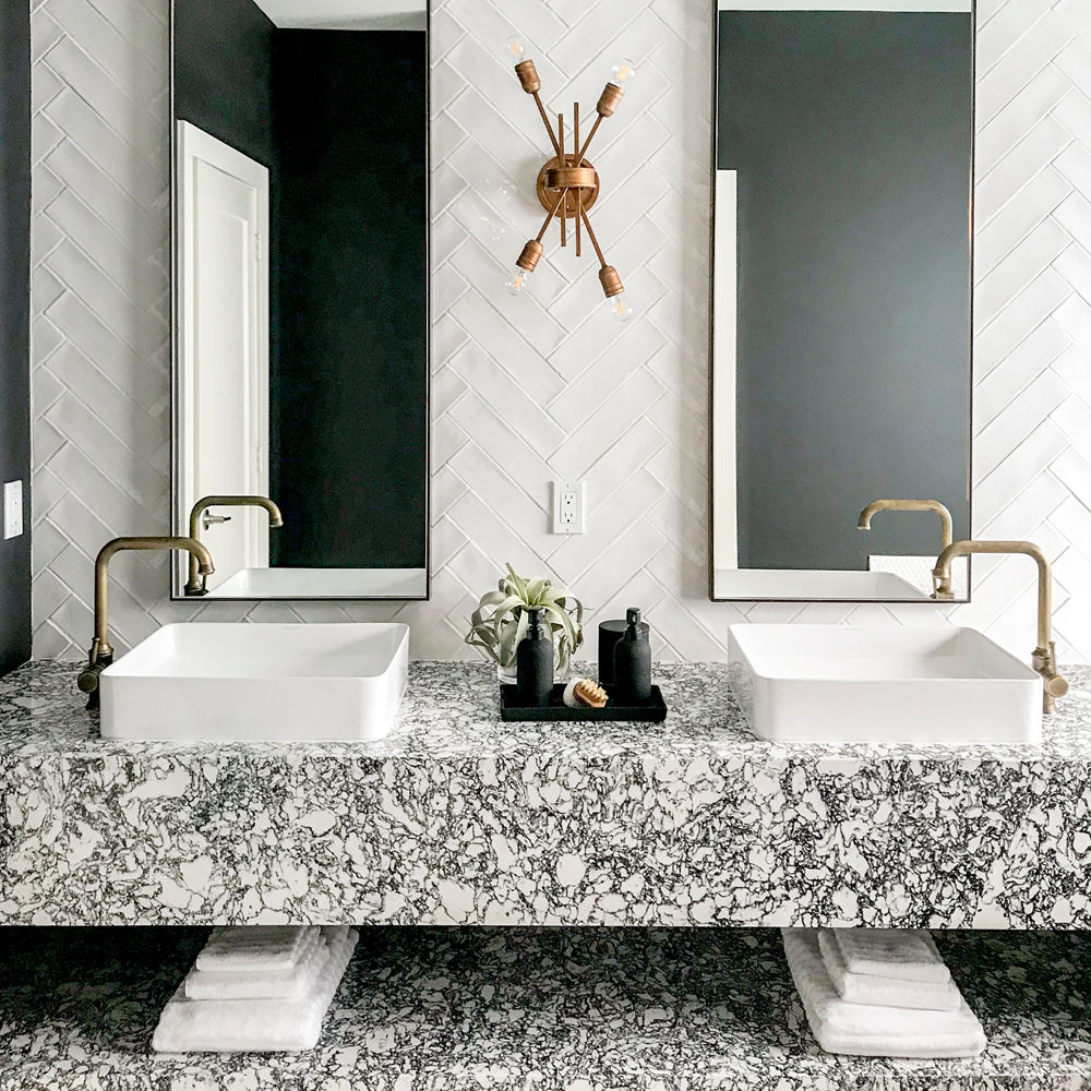 a bathroom with a floating shelf vanity and shelf made from black and white speckled quartz, with two vessel sinks with gold faucets, two long, veritcal mirrors, and white herringbone tile backsplash.