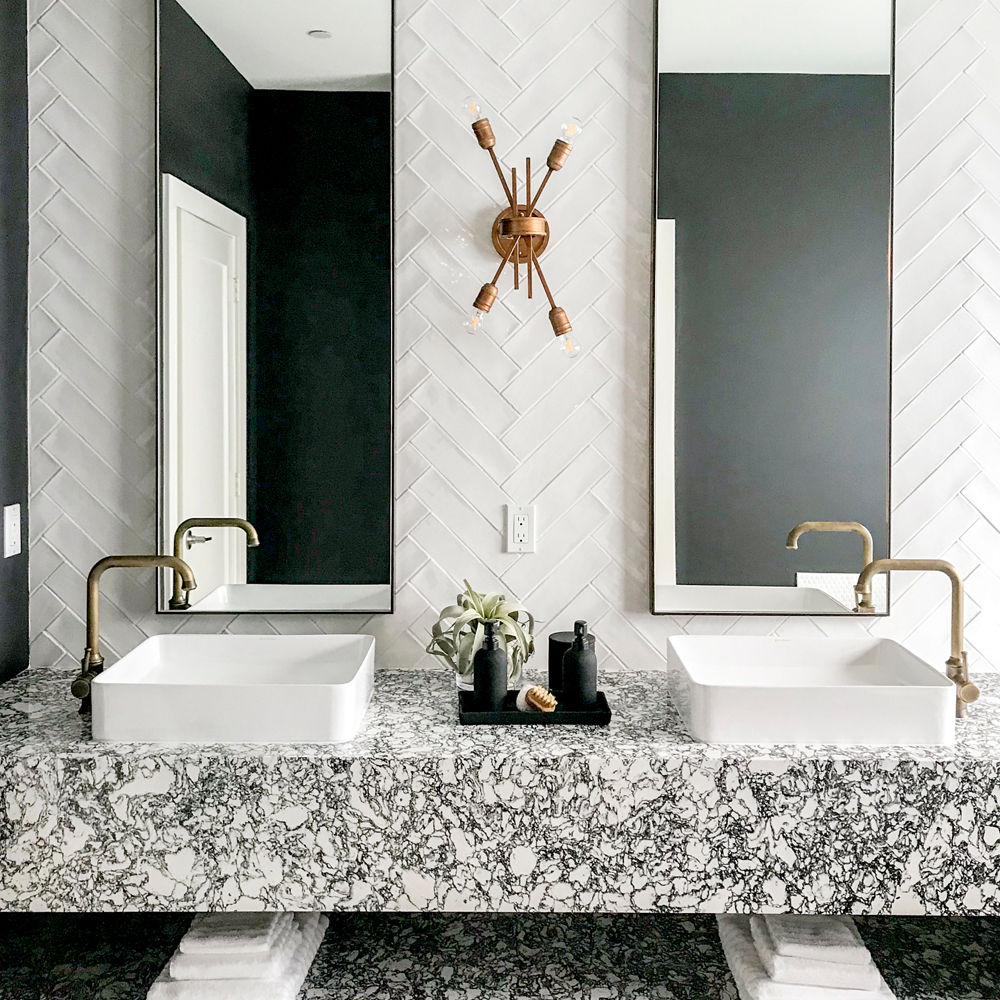 a bathroom with a floating shelf vanity and shelf made from black and white speckled quartz, with two vessel sinks with gold faucets, two long, veritcal mirrors, and white herringbone tile backsplash.