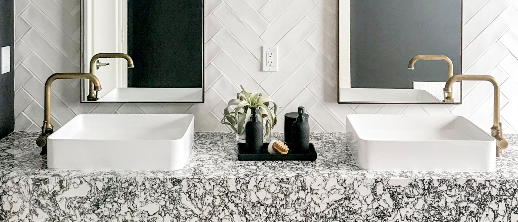 a bathroom with a floating shelf vanity and shelf made from black and white speckled quartz, with two vessel sinks with gold faucets, two long, veritcal mirrors, and white herringbone tile backsplash.