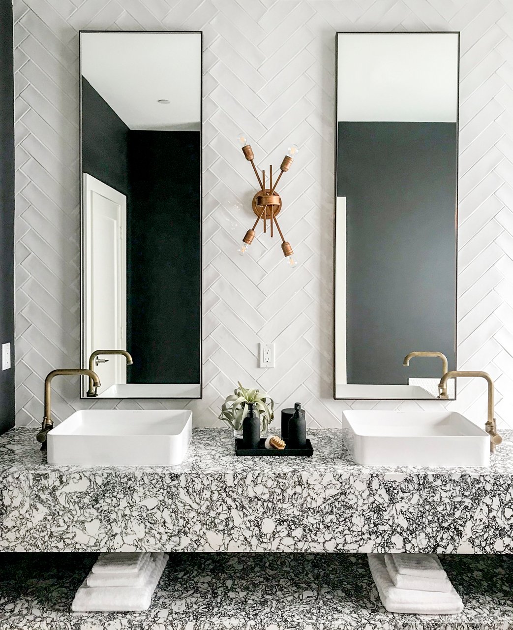 a bathroom with a floating shelf vanity and shelf made from black and white speckled quartz, with two vessel sinks with gold faucets, two long, veritcal mirrors, and white herringbone tile backsplash.