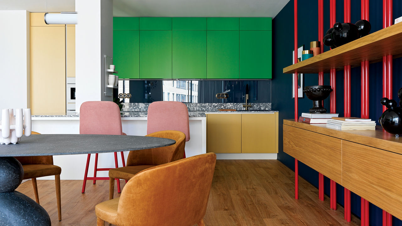 a bold kitchen with green upper cabinets, yellow lower cabinets, and black and white speckled countertops and backsplash, with shelving to the right with red pipes behind it, and a dining table with leather seats