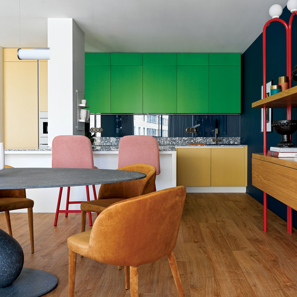 a bold kitchen with green upper cabinets, yellow lower cabinets, and black and white speckled countertops and backsplash, with shelving to the right with red pipes behind it, and a dining table with leather seats
