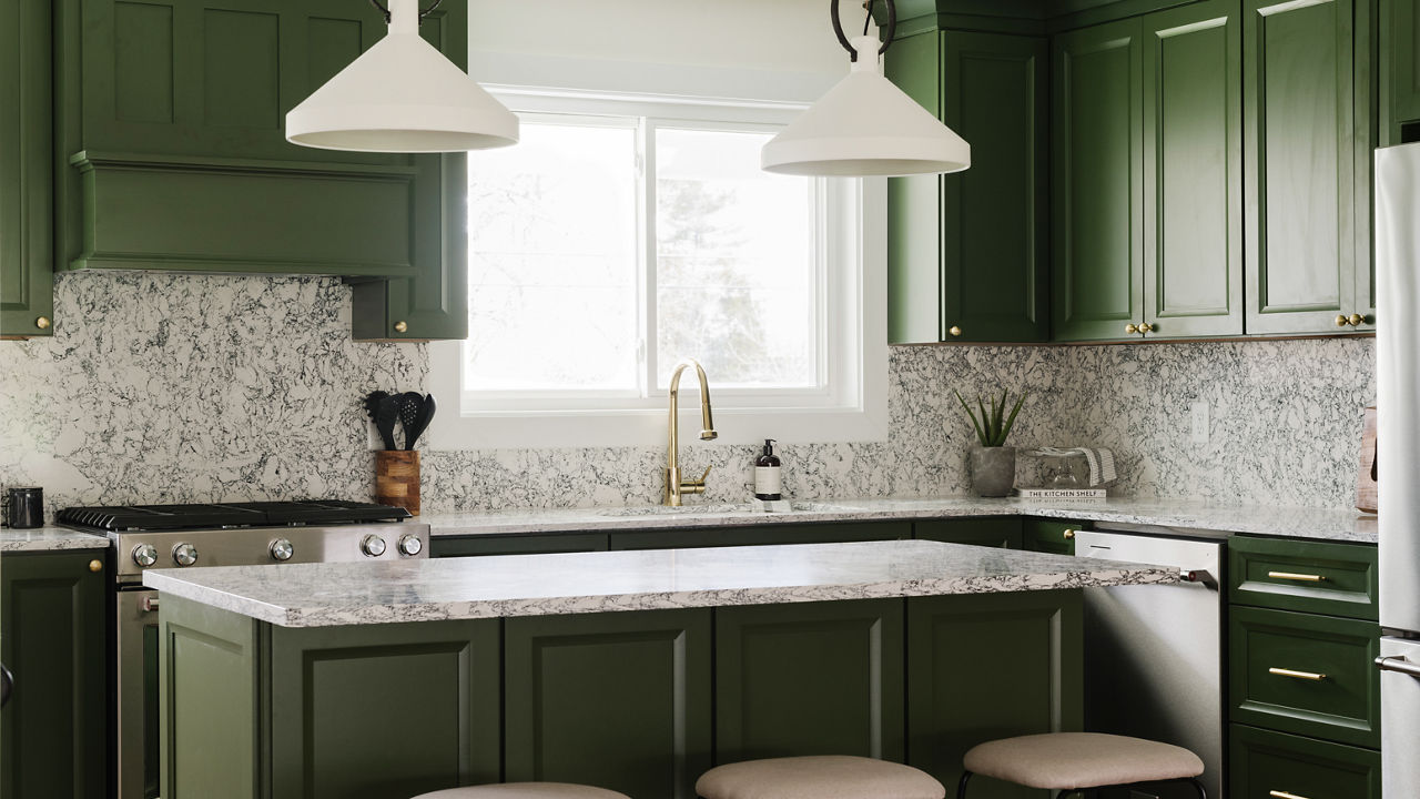 a mossy green kitchen with green cabinets topped with black and white speckled quartz countertops and backsplash, with three cream colored barstools and overhead pendant lighting