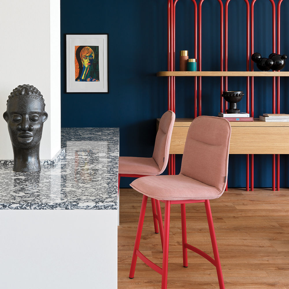 a kitchen with white and black speckled quartz countertops, colorful red and pink barstools, a navy blue wall, and wooden flooring.