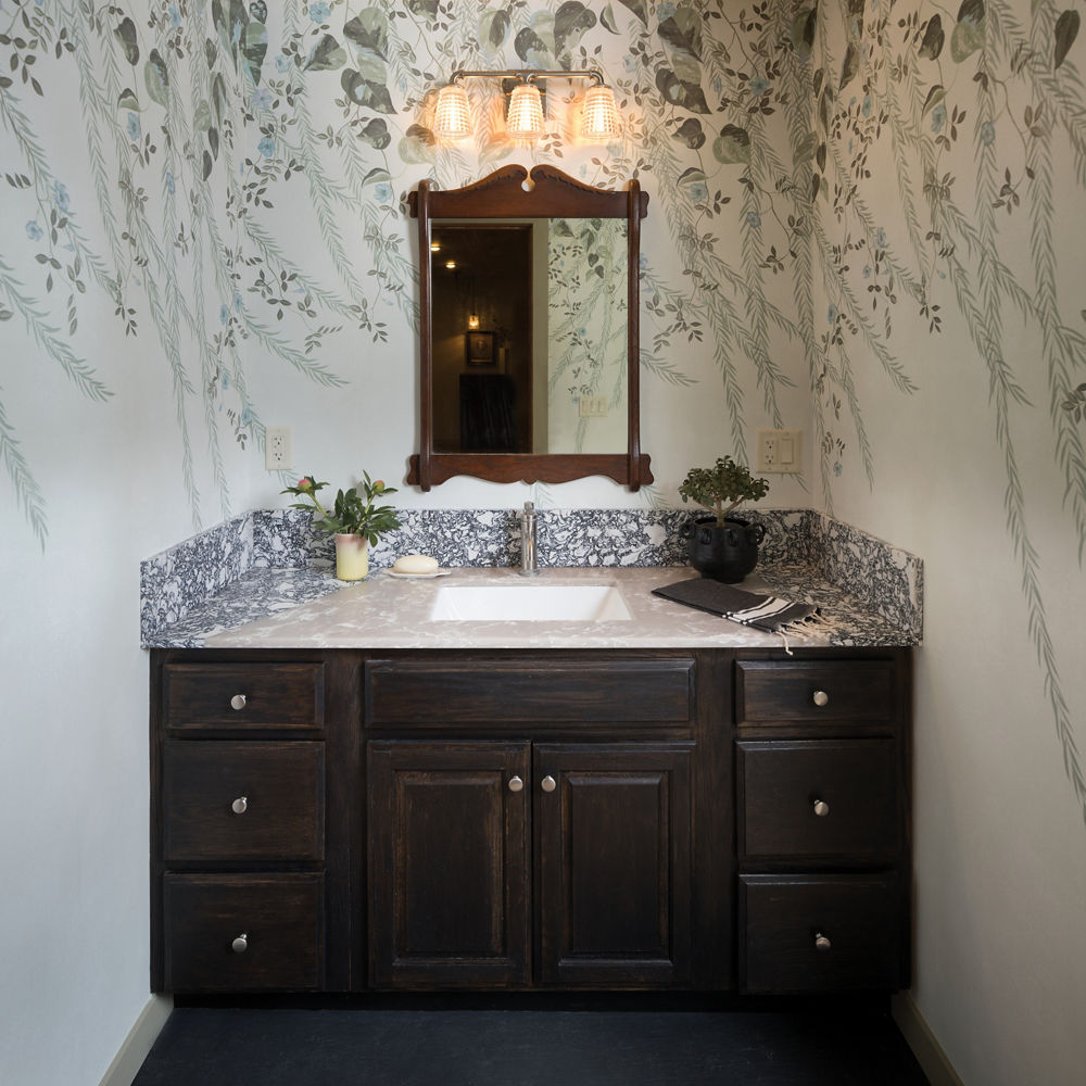 A bathroom with floral wall paper, black wooden vanity, black and white quartz countertop, and a antique mirror.