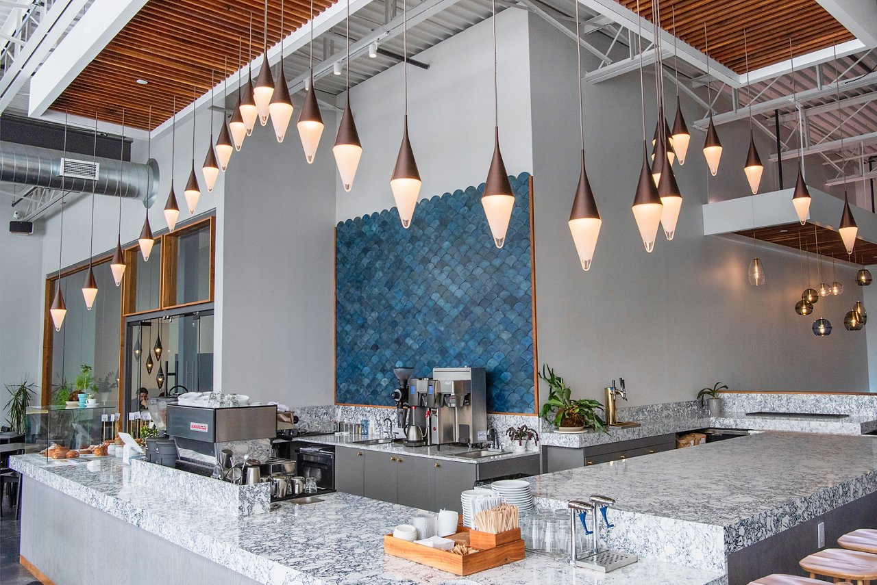 a cafe with an espresso machine, organic pendant lighting, countertops made from white and black quartz, a unique blue backsplash, and lots of natural light. 