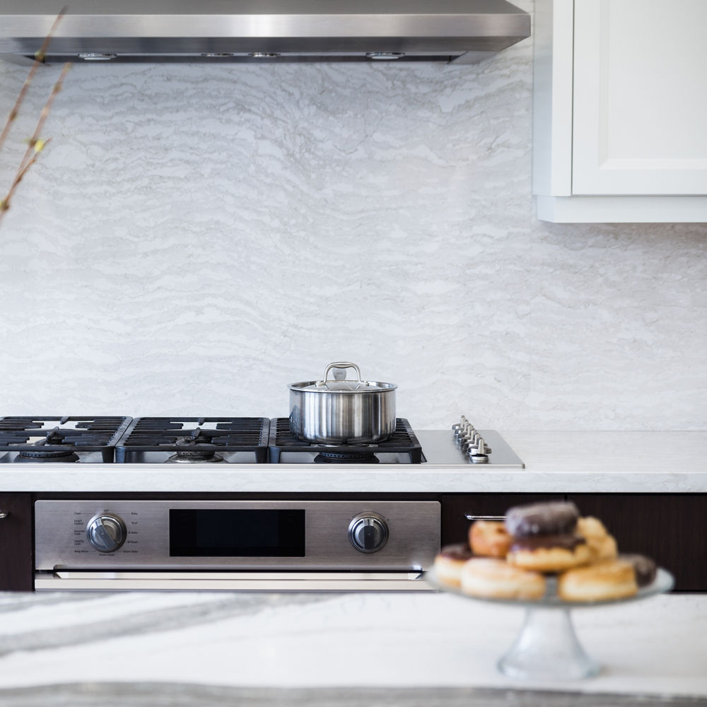 A kitchen with an island topped with gray and white veined quartz countertops with a range in the background and a quartz backsplash.