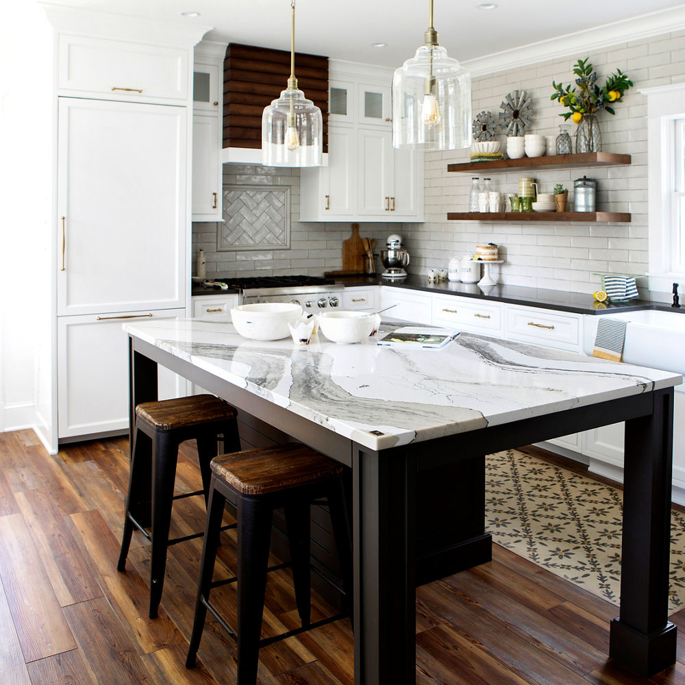 A traditional kitchen with an open black island topped with gray and white veined quartz countertops with two barstools and pendant lighting, the rest of the kitchen has white cabinets and open wooden upper cabinets. 