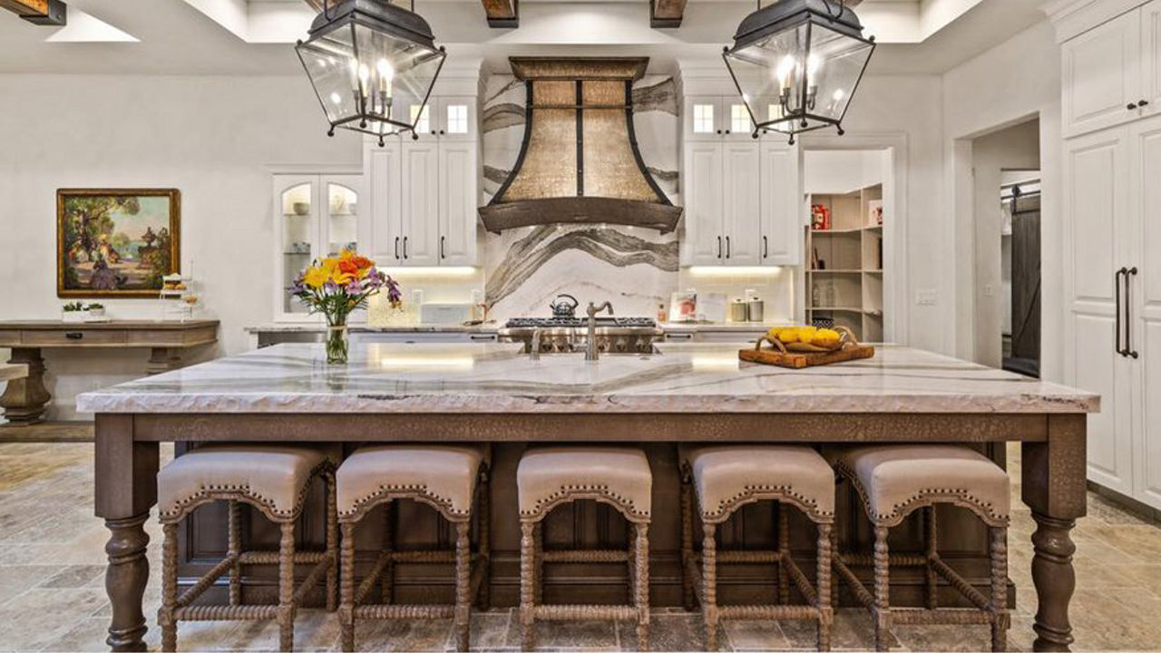 A traditional kitchen with white cabinets, wooden beams on the ceiling, large central island with barstools and quartz countertops with matching quartz backsplash, bold brown and tan hood, and pendant lighting. 