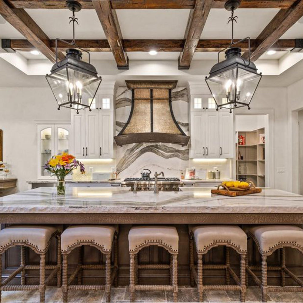 A traditional kitchen with white cabinets, wooden beams on the ceiling, large central island with barstools and quartz countertops with matching quartz backsplash, bold brown and tan hood, and pendant lighting. 