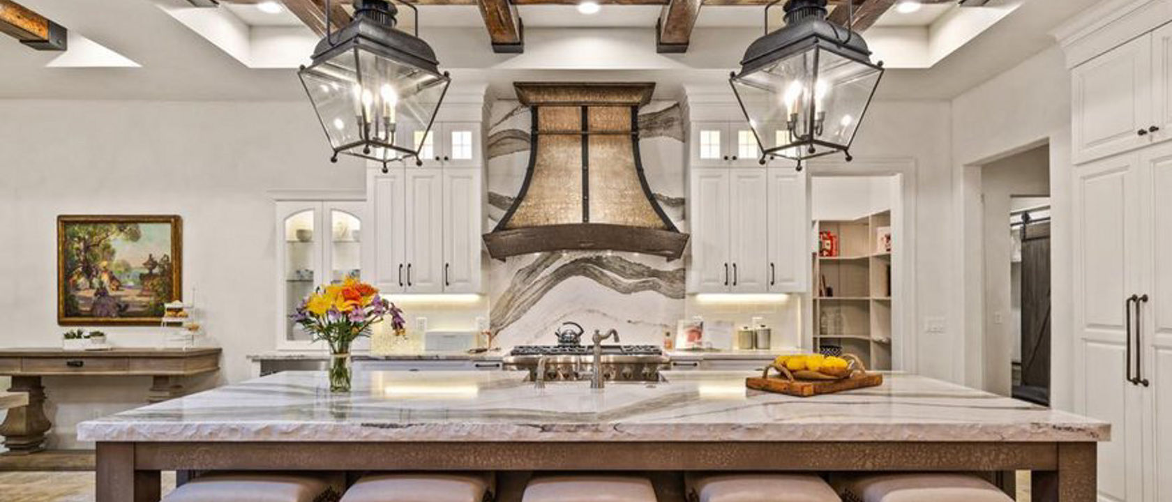 A traditional kitchen with white cabinets, wooden beams on the ceiling, large central island with barstools and quartz countertops with matching quartz backsplash, bold brown and tan hood, and pendant lighting. 