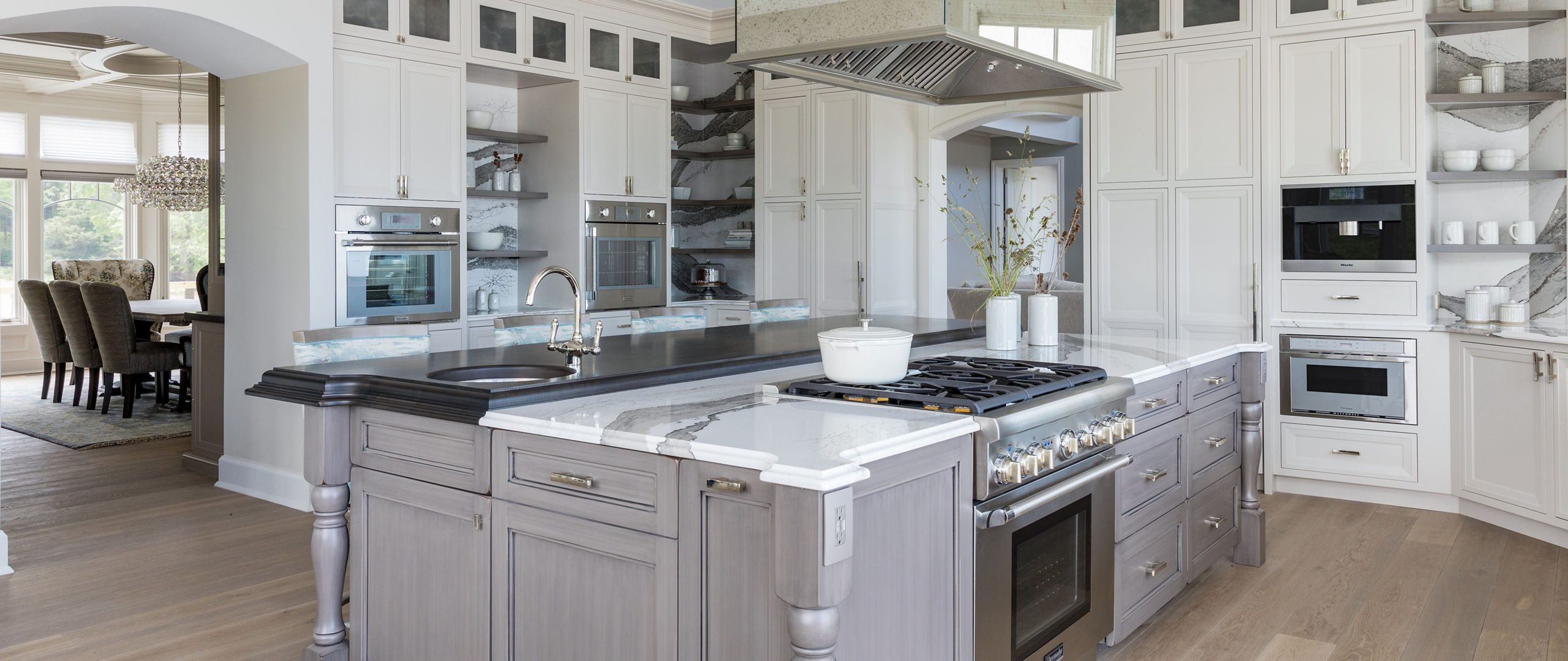 a large traditional kitchen with white upper and lower cabinets, some open shelving, white and gray veined quartz backsplash and countertops, a gray wooden island topped with matching quartz countertops with a built in stove in the oven. 