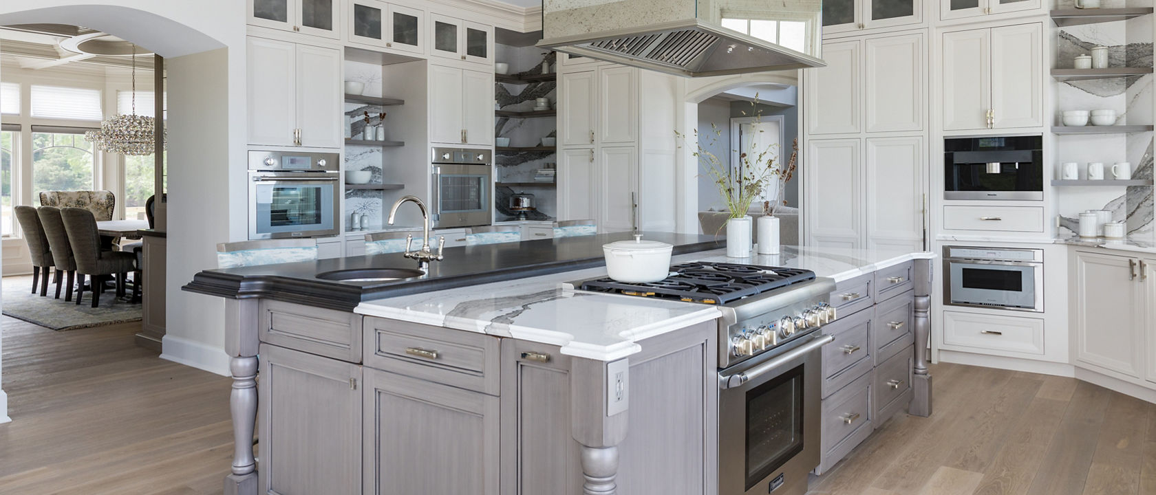 a large traditional kitchen with white upper and lower cabinets, some open shelving, white and gray veined quartz backsplash and countertops, a gray wooden island topped with matching quartz countertops with a built in stove in the oven. 