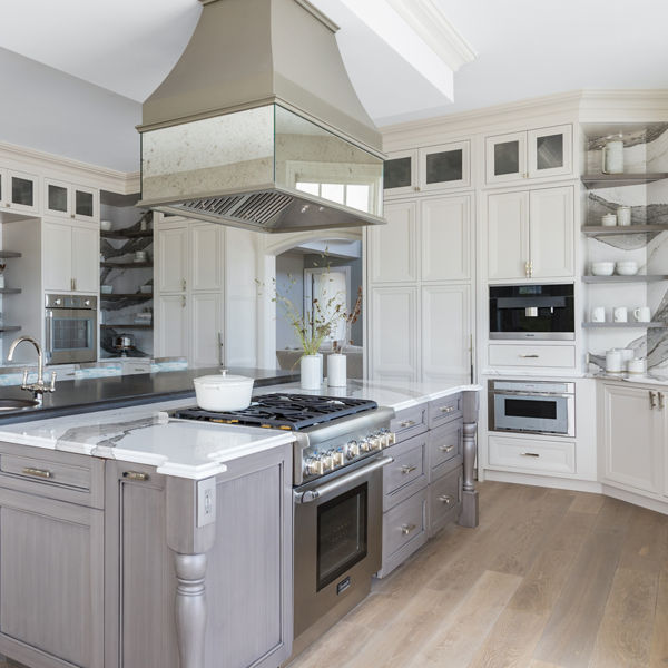 a large traditional kitchen with white upper and lower cabinets, some open shelving, white and gray veined quartz backsplash and countertops, a gray wooden island topped with matching quartz countertops with a built in stove in the oven. 