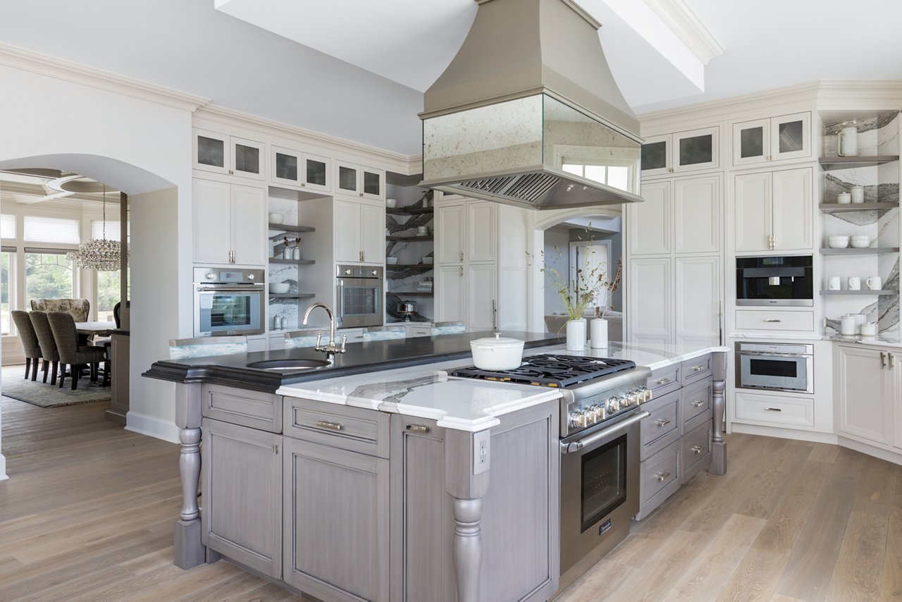 a large traditional kitchen with white upper and lower cabinets, some open shelving, white and gray veined quartz backsplash and countertops, a gray wooden island topped with matching quartz countertops with a built in stove in the oven. 