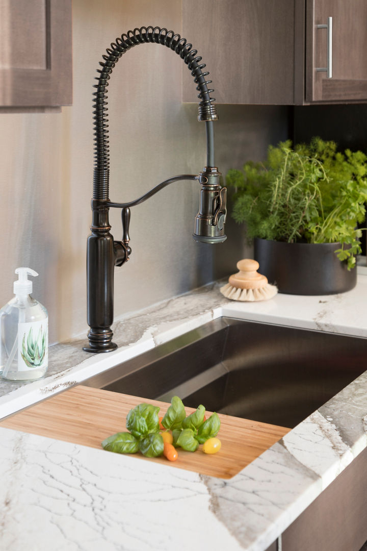 A close up of a sink with white and gray veined quartz countertops, brown cabinets and a black pullout faucet with a cutting board built into the left side
