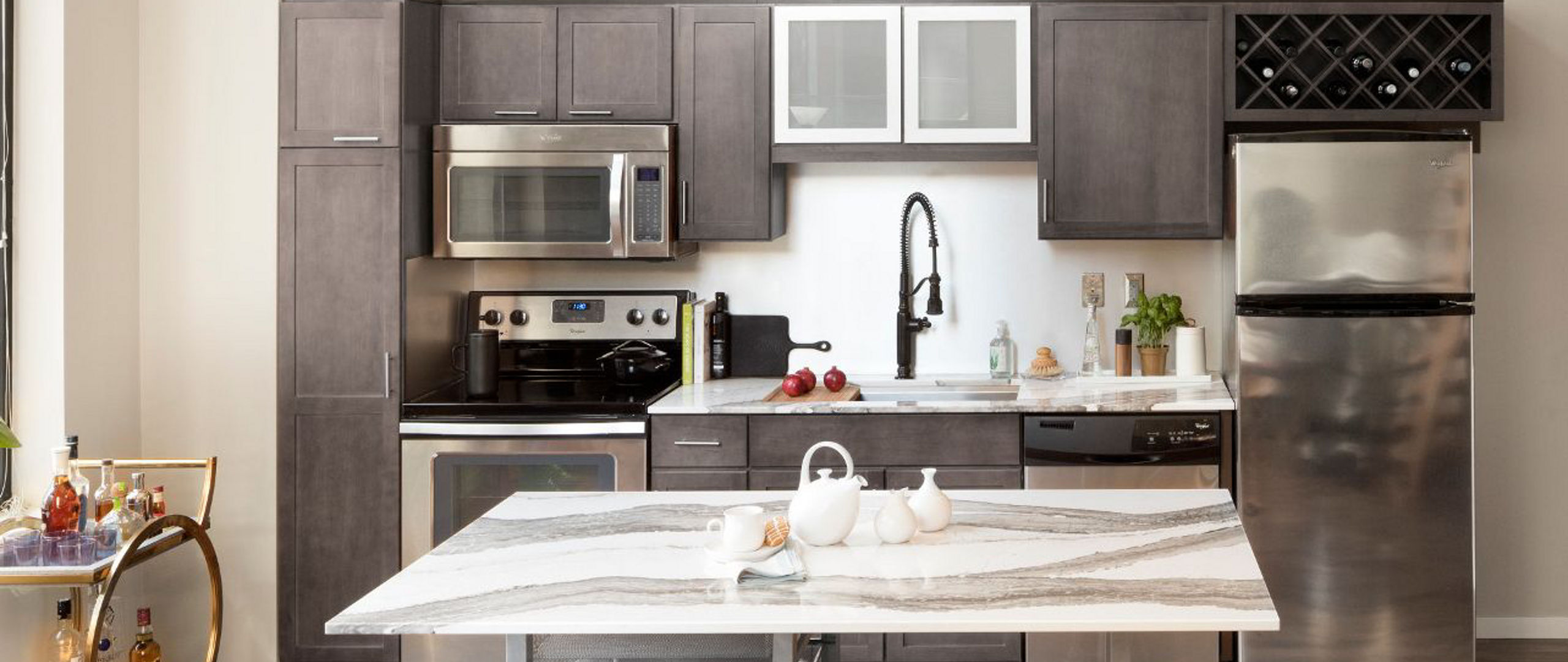 A gorgeous kitchen with light gray-brown cabinets, white quartz countertops, a center island topped with white quartz, and modern appliances. 