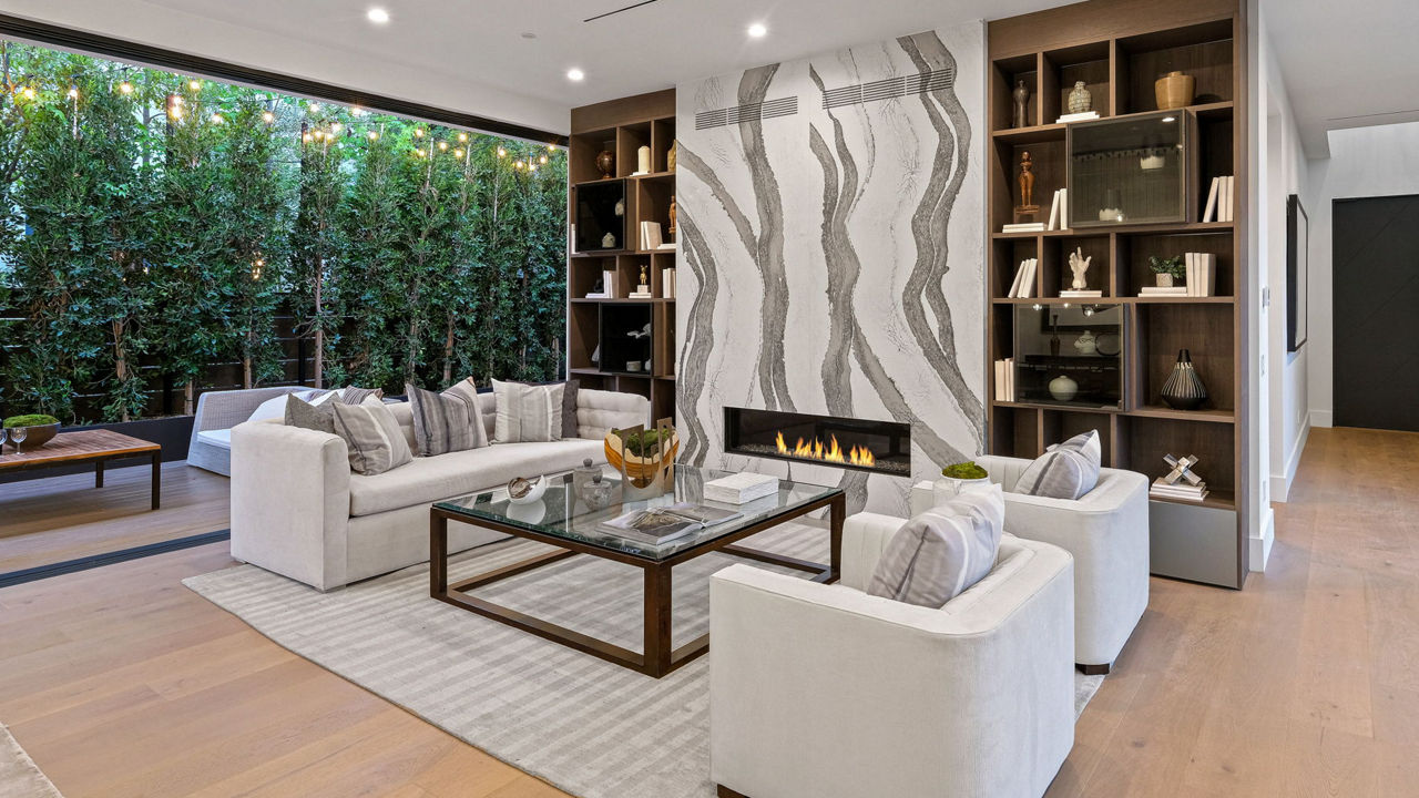 A stunning living room with a white and gray veined fireplace, built in bookshelves, white seating, and a large window.