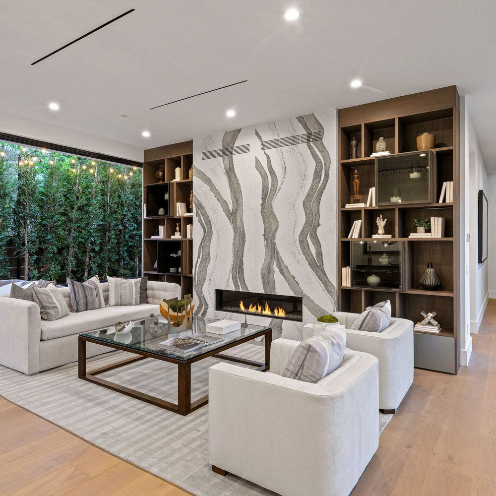 A stunning living room with a white and gray veined fireplace, built in bookshelves, white seating, and a large window.