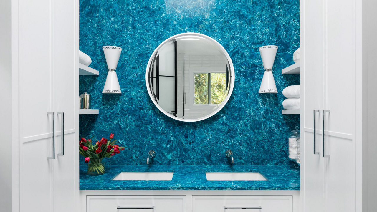 A bold bathroom with blue quartz countertops and backsplash with a white, circular mirror, two white sconces, two sinks, and white cabinets.