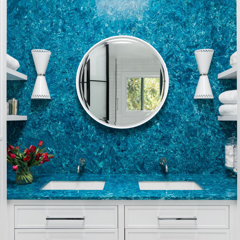 A bold bathroom with blue quartz countertops and backsplash with a white, circular mirror, two white sconces, two sinks, and white cabinets.