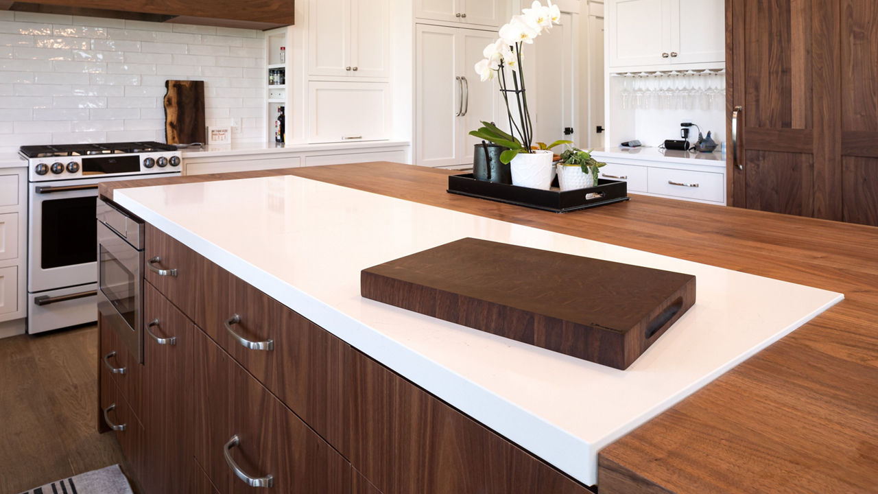 a traditional kitchen with oak island cabinets topped with butcher block countertops with a slab of white quartz for accent, while the rest of the kitchen has white cabinets, white tile backsplash, and wooden accents