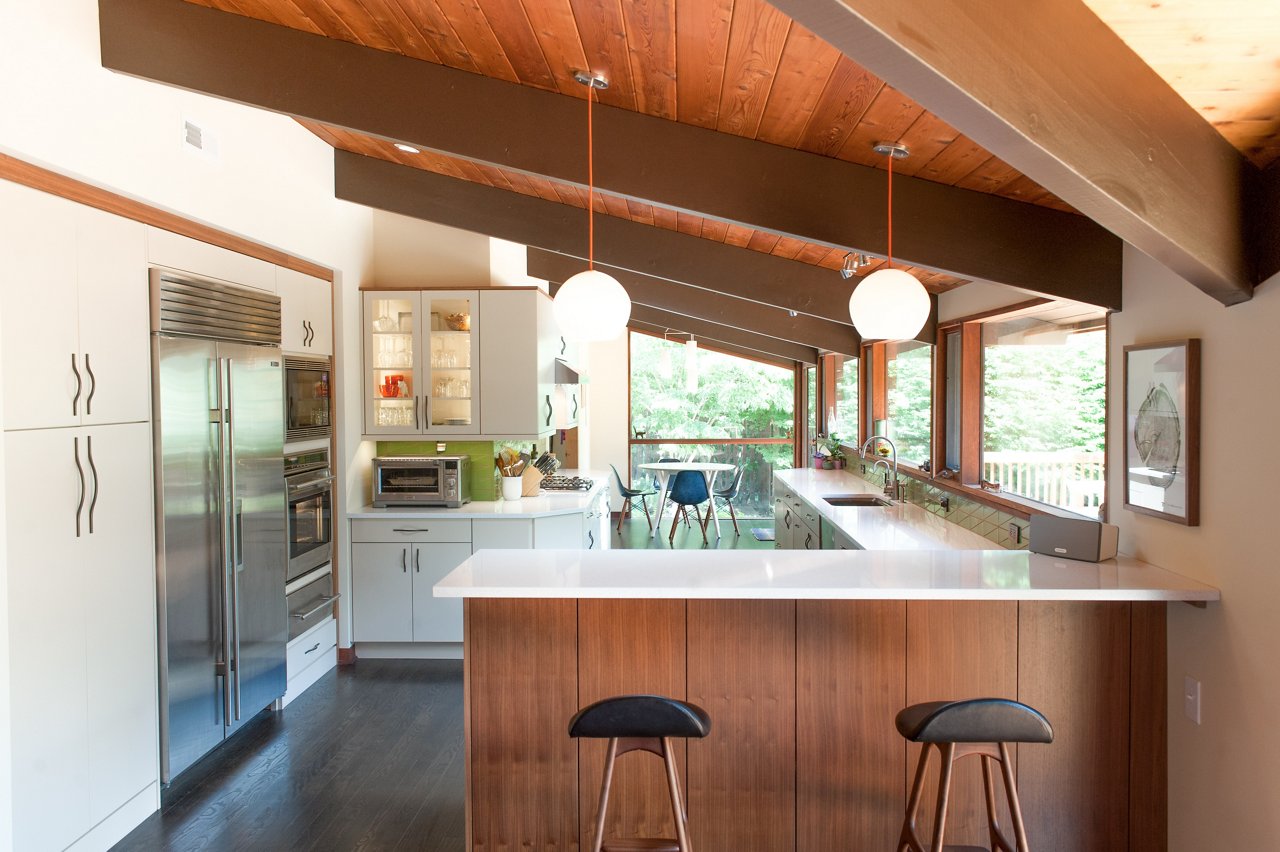 A warm kitchen with oak cabinets, half vaulted wooden ceilings, stainless steel appliances, and Snowdon White quartz countertops