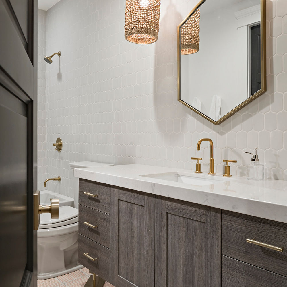 A bathroom with dark oak cabinets, white quartz countertops, brass faucet, hexagonal mirror with gold trim, two overhead pendant lights, and brown and white tile floors