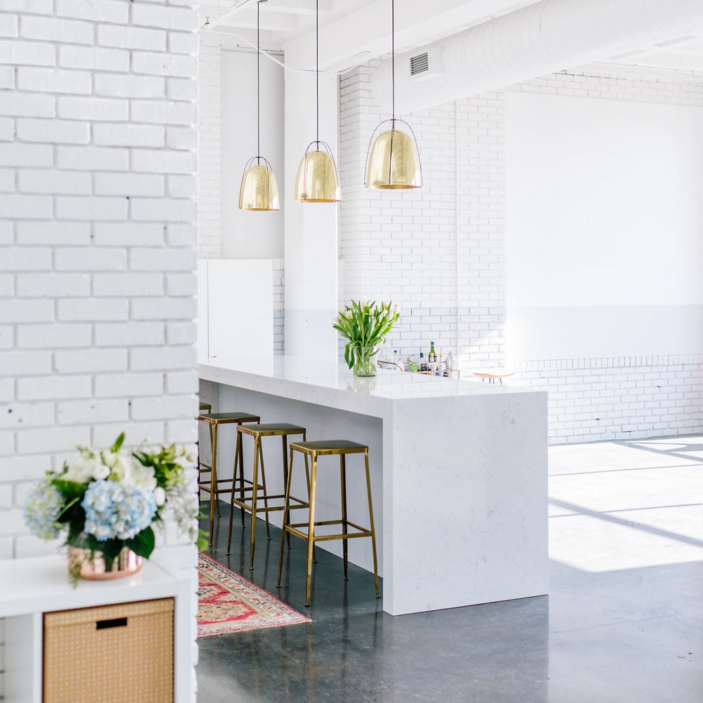 a clean kitchen with exposed white bricks, a double waterfall island made from white quartz, gold barstools and concrete floors.