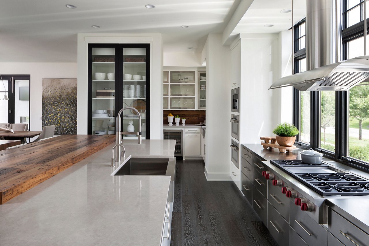 A modern kitchen with gray quartz countertops, gray cabinets, a modern range and hood, dark hardwood floors, and large windows letting in natural light.