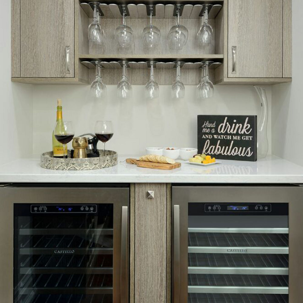 a home dry bar with two mini fridges, white quartz countertops, and shelves built to hang cocktail glasses from. 