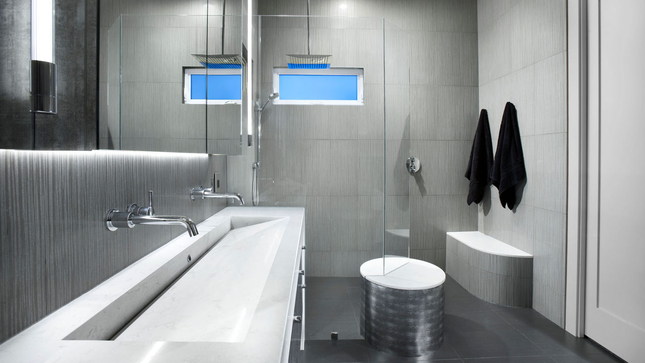 an industrial-modern bathroom with a gray vanity topped with white quartz countertops and integrated sink with a glass shower to the right of the vanity, two black robes hanging on the wall, and dark black tiles on the floor