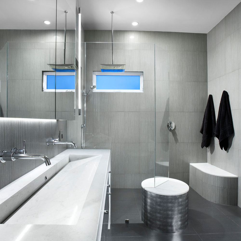 an industrial-modern bathroom with a gray vanity topped with white quartz countertops and integrated sink with a glass shower to the right of the vanity, two black robes hanging on the wall, and dark black tiles on the floor