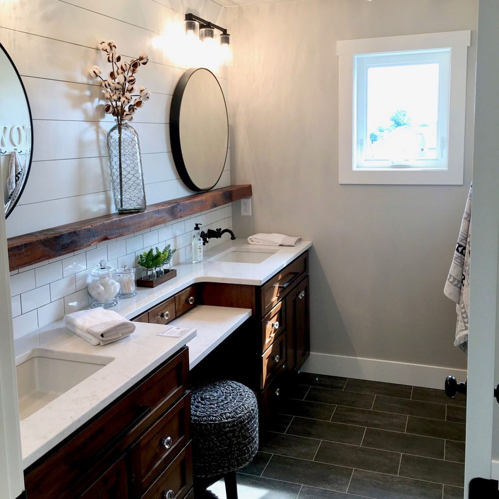 A bathroom with dark oak cabinets, white quartz countertops, white subway tile, dark tile floors, shiplap walls, two rounded mirrors, and a pouf vanity in the middle