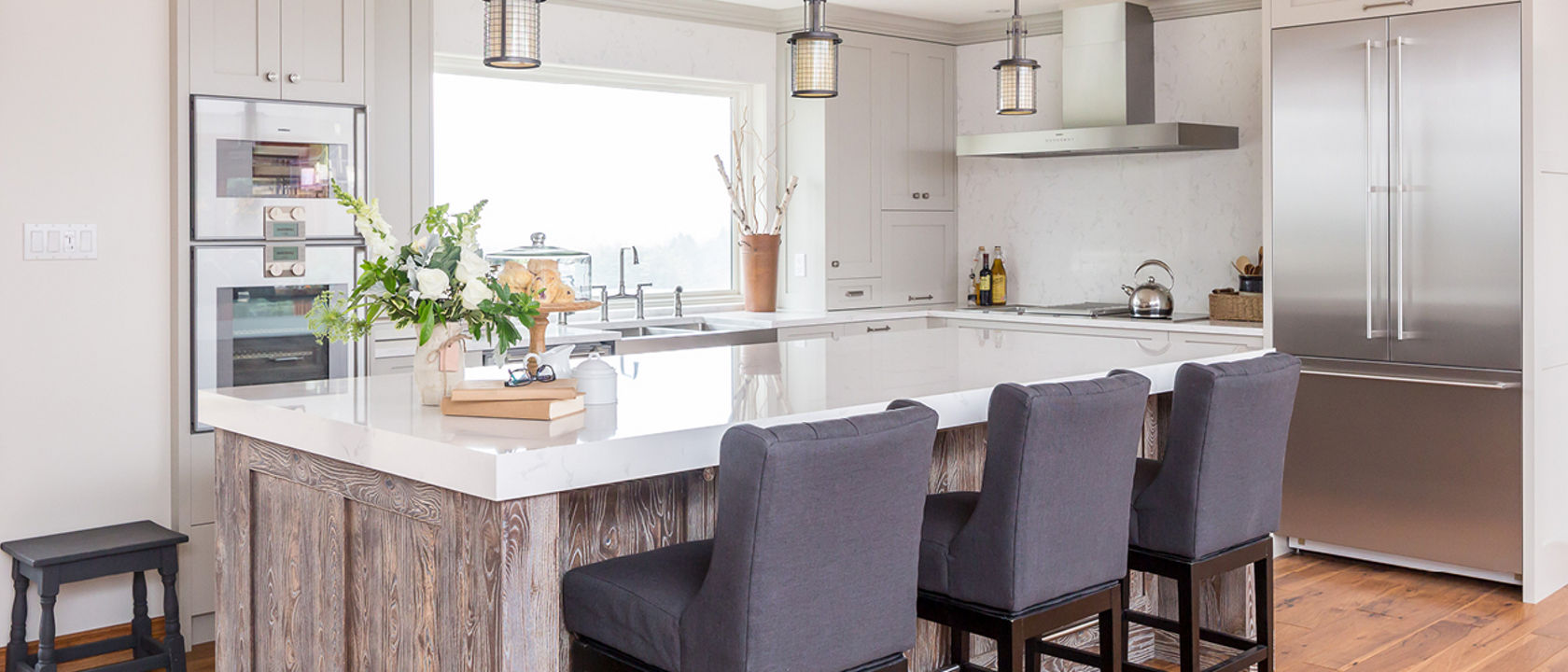 A gorgeous kitchen with a wooden island topped with Cambria Quartz Torquay countertops, three blue barstools, large window over the sink, and modern range.