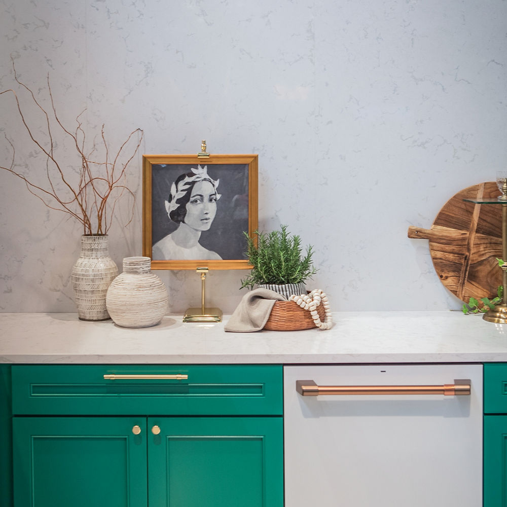 a kitchen with emerald green lower cabinets, a white dishwasher with gold handle, white quartz countertops, with a picture, vase, and plant sitting on top of the counters