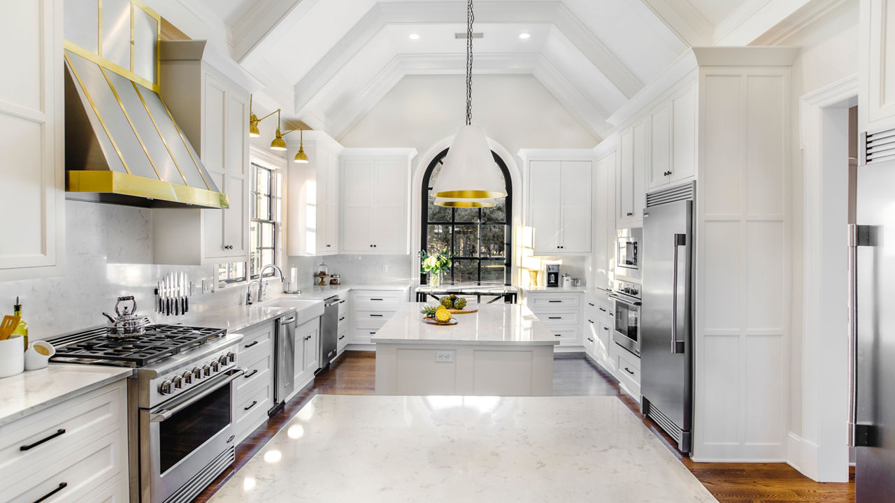 A beautiful white kitchen with two islands topped with white quartz countertops, stainless steel appliances, white cabinets, black and gold accents throughout, and vaulted ceilings to create a spacious feel.