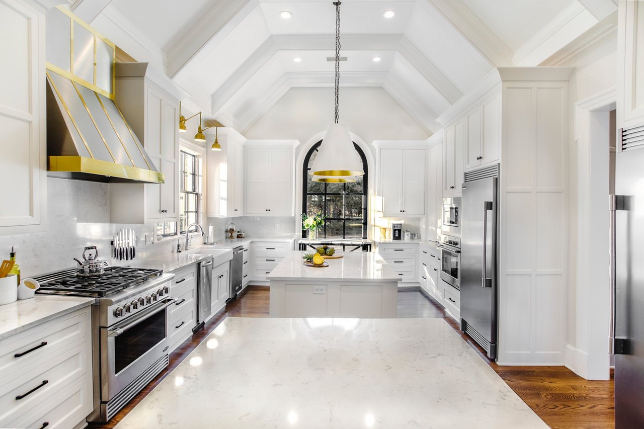 A beautiful white kitchen with two islands topped with white quartz countertops, stainless steel appliances, white cabinets, black and gold accents throughout, and vaulted ceilings to create a spacious feel.