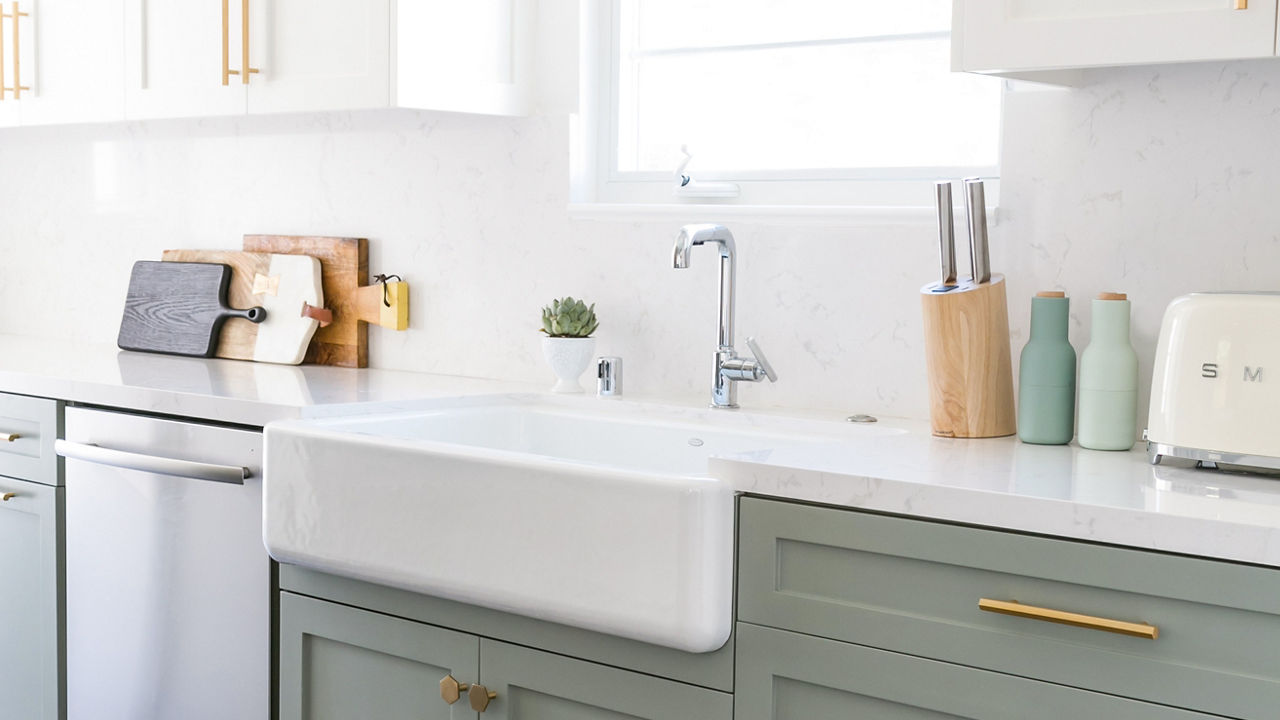 a kitchen with light green lower cabinets, white upper cabinets, both with gold handles, a farmhouse sink with silver faucet, and miscellaneous kitchen appliances and supplies sitting on the counters.