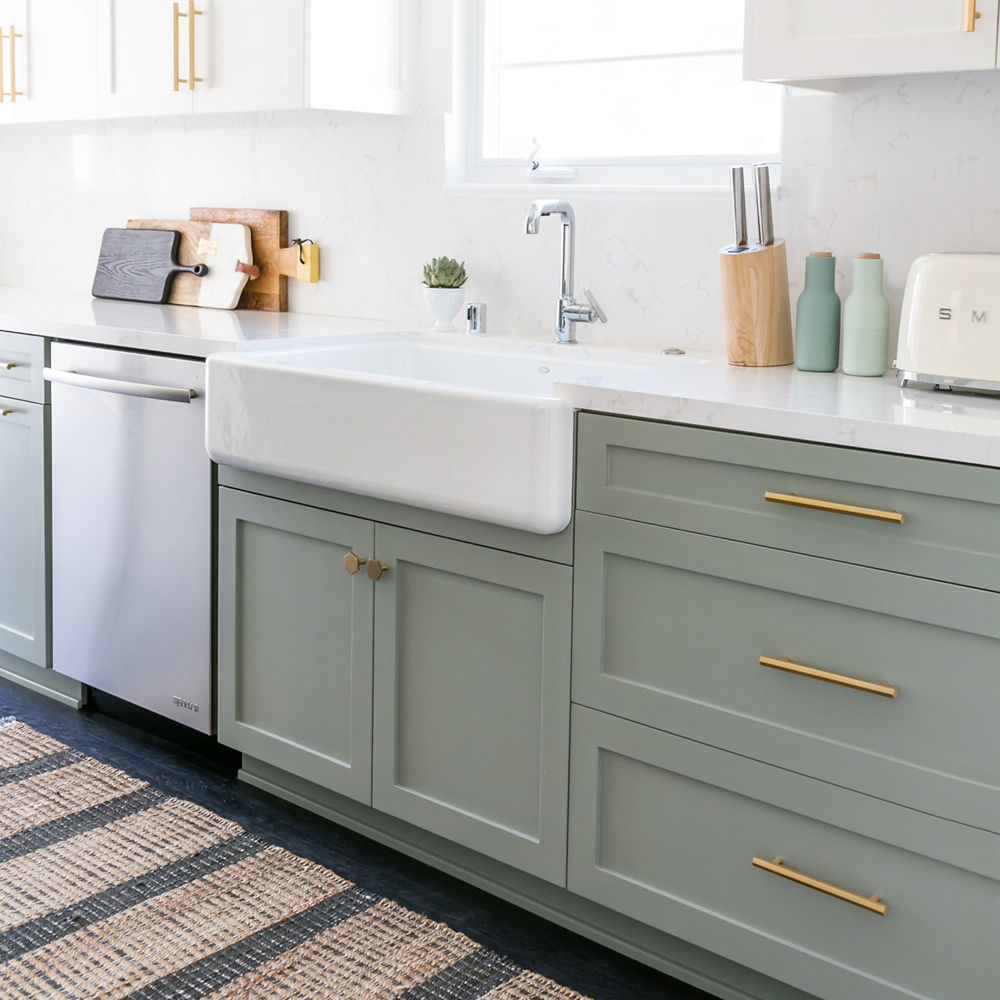 a kitchen with light green lower cabinets, white upper cabinets, both with gold handles, a farmhouse sink with silver faucet, and miscellaneous kitchen appliances and supplies sitting on the counters.
