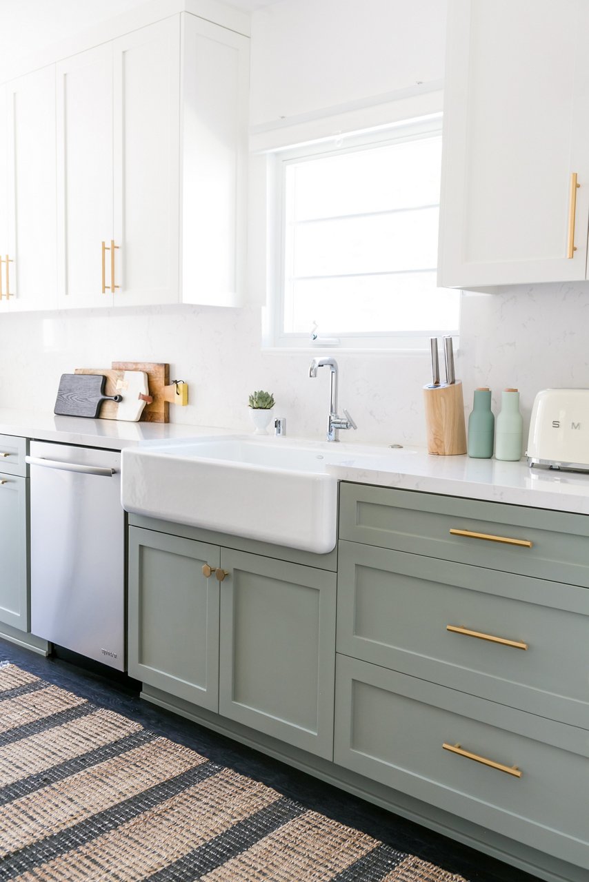 a kitchen with light green lower cabinets, white upper cabinets, both with gold handles, a farmhouse sink with silver faucet, and miscellaneous kitchen appliances and supplies sitting on the counters.