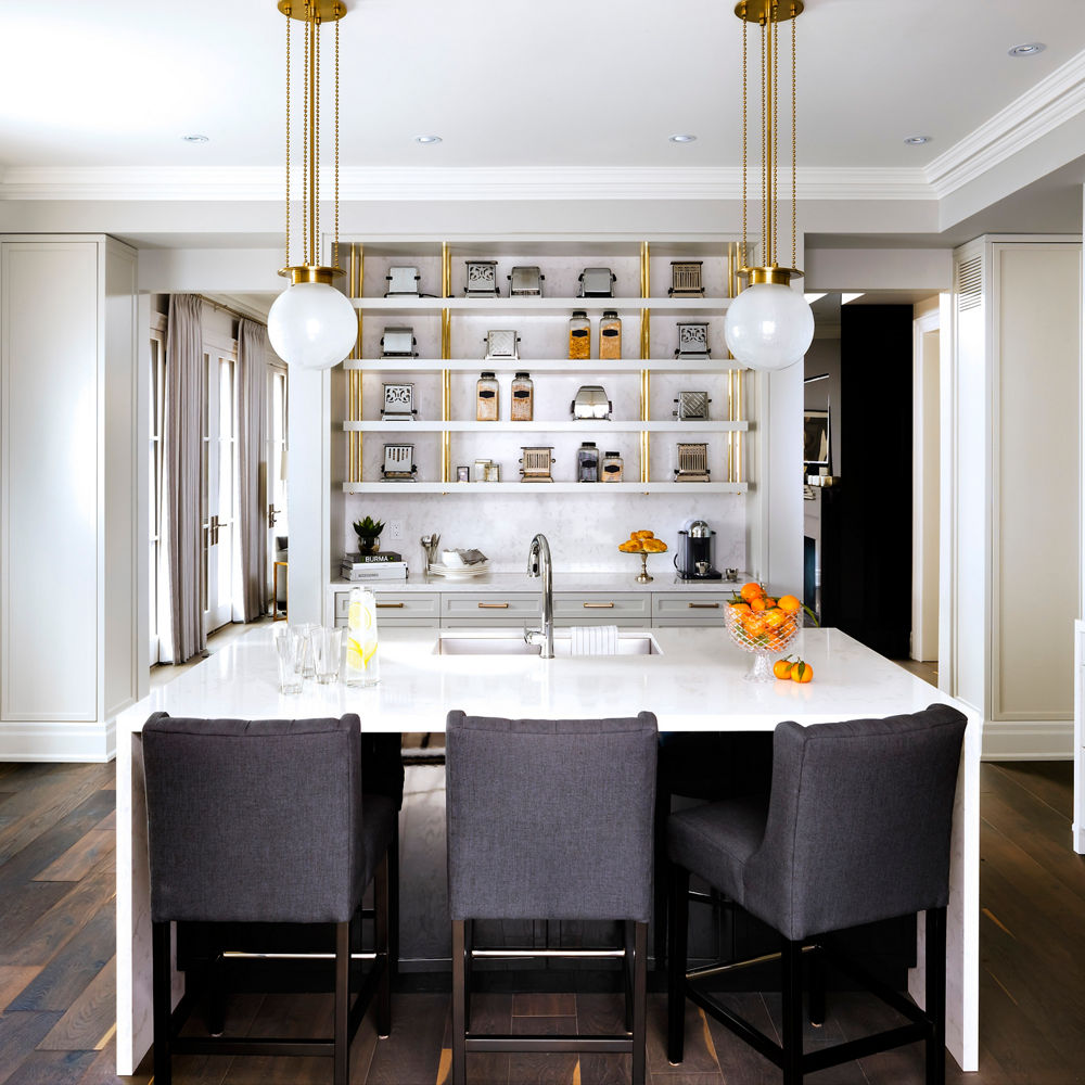 A kitchen island topped with a double waterfall-edge quartz countertop with three barstools and a built-in sink with overhead lighting and wooden flooring.