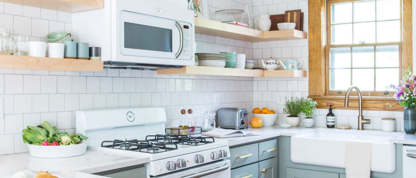 a galley kitchen with light green cabinets, farmhouse sink, open wooden shelving, white quartz countertops, a wooden widow, and subway tile.