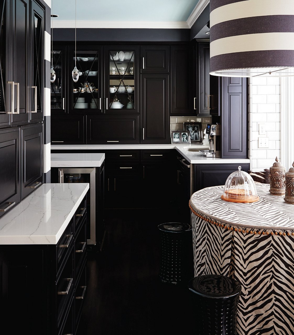 a dark and sleek modern kitchen with black cabinets, white quartz countertops, a dining table with a zebra pattern table cloth and a striped chandelier above it.