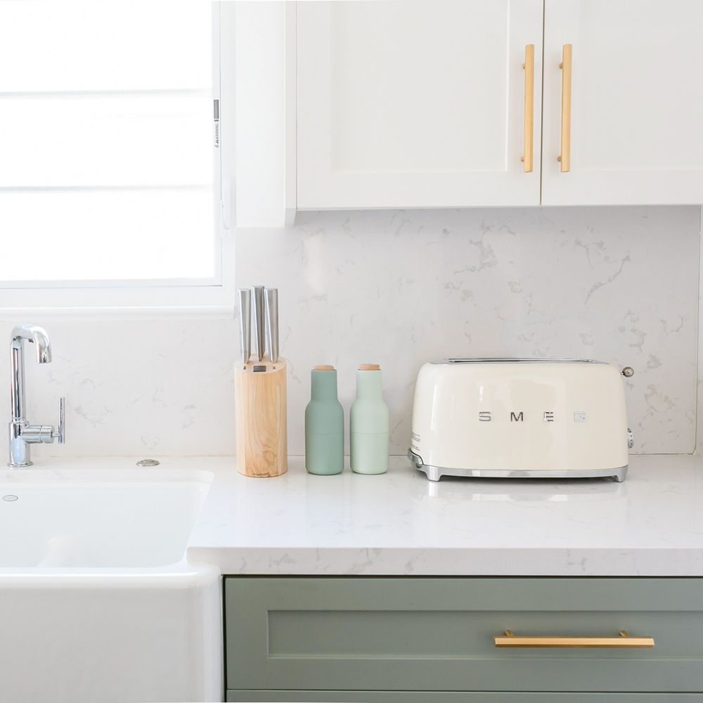 a kitchen with light green lower cabinets, white upper cabinets, both with gold handles, a farmhouse sink with silver faucet, and a Smeg toaster sitting on the countertop.