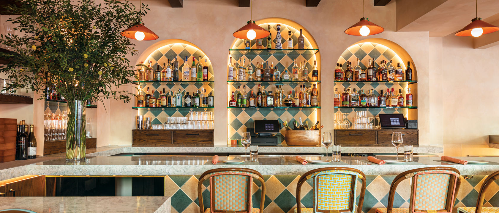a bold kitchen bar with a teal and cream diamond pattern on the front face of bar and backsplash, beige quartz countertops, wicker and wood barstools, orange pendant lighting and giant plant on the bar. 