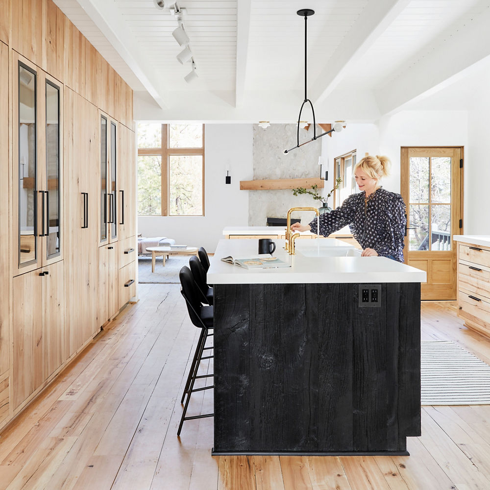 Emily Henderson in her mountain house kitchen with oak floors and cabinets, a black center island topped with white quartz countertops, and paneled ceilings.