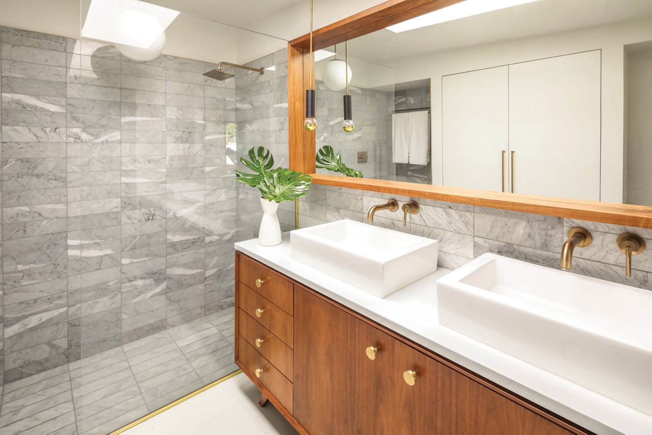 A mid-century modern bathroom with oak cabinets, white quartz countertops, a large mirror framed with wood, and a gorgeous tiled shower. 