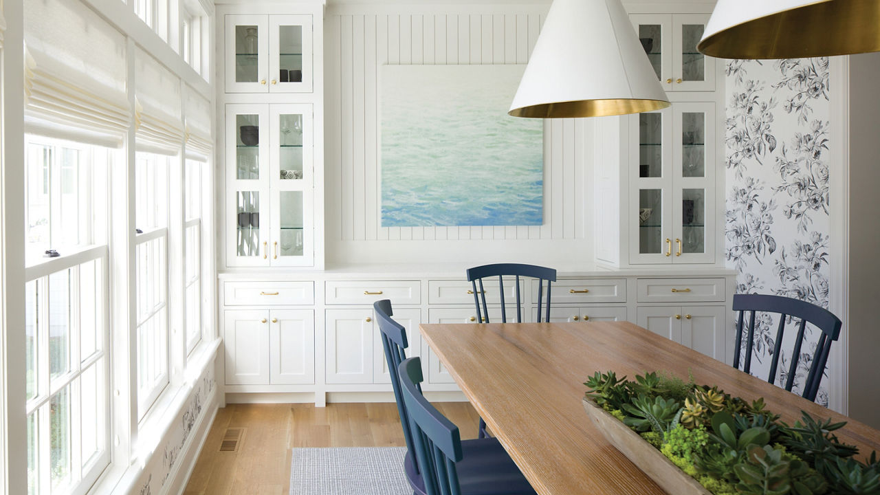 A coastal inspired dining room with blue chairs, white hutch topped with white quartz, a sea-inspired painting, and wooden flooring.
